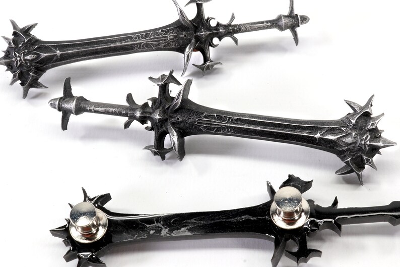 May include: Three silver-colored sword pins with intricate details and a black finish. The pins have a sharp point and a decorative design.