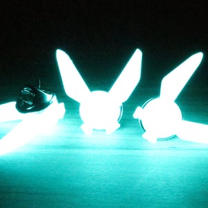 Peut inclure: Trois &eacute;pingles qui brillent dans le noir en forme d'oiseau stylis&eacute; avec les ailes d&eacute;ploy&eacute;es. Les &eacute;pingles sont de couleur turquoise brillante et se trouvent sur une surface sombre.