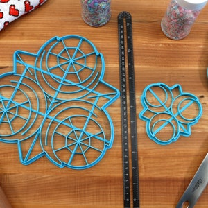 May include: A set of three blue plastic cookie cutters, one large and two smaller, on a wooden surface. The large cutter is shaped like a snowflake with a circular design. The smaller cutters are shaped like circles with a line across the center. A ruler, a rolling pin, and a blue spatula are also visible in the image.