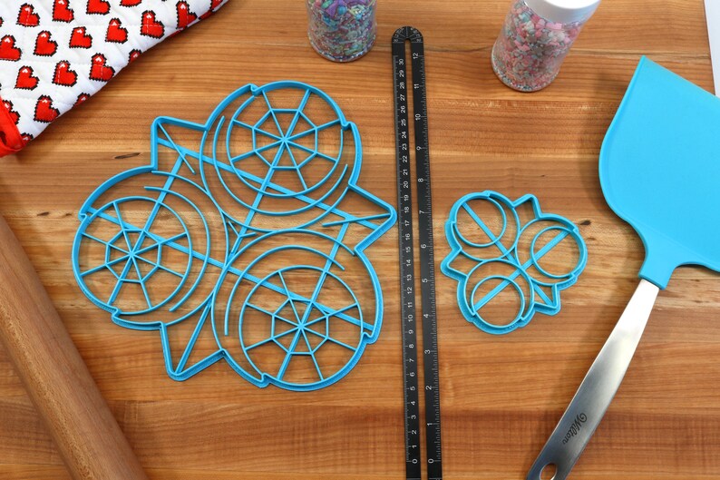 May include: A set of three blue plastic cookie cutters, one large and two smaller, on a wooden surface. The large cutter is shaped like a snowflake with a circular design. The smaller cutters are shaped like circles with a line across the center. A ruler, a rolling pin, and a blue spatula are also visible in the image.