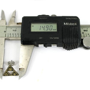 May include: A silver-colored metal earring with a triangular design featuring a stylized symbol. The earring is being measured with a digital caliper, which reads 14.90 mm.