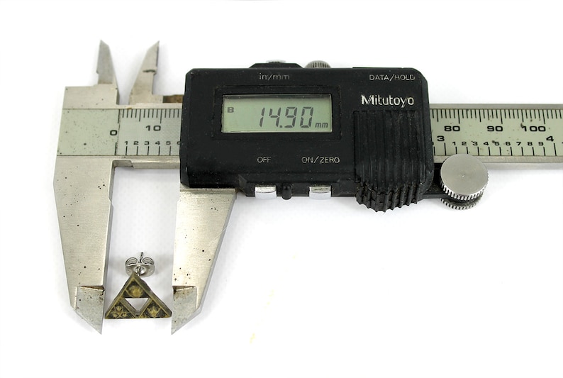 May include: A silver-colored metal earring with a triangular design featuring a stylized symbol. The earring is being measured with a digital caliper, which reads 14.90 mm.