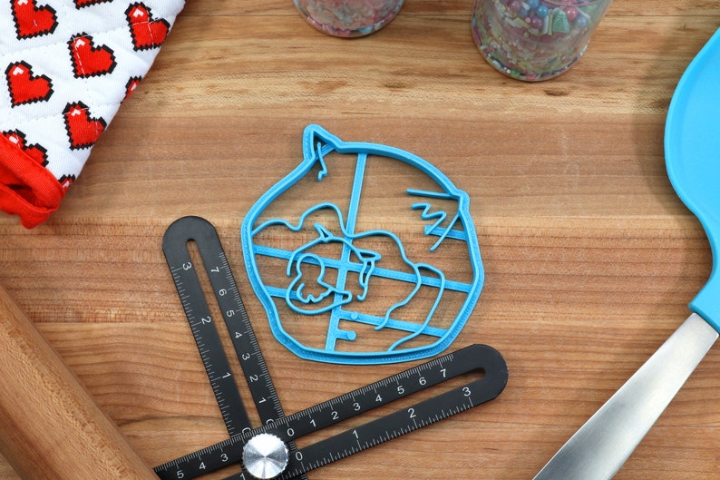 May include: A blue plastic cookie cutter shaped like a cartoon animal with a large head and a small body. The cutter has a detailed design with lines and curves.