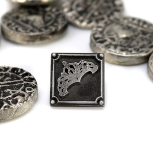 May include: Close-up of antique silver-colored coins and a square token. The token features a decorative design within a square frame. The coins have various embossed patterns. The items are arranged on a white surface, creating a contrast with the silver tones.