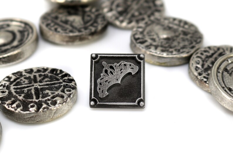 May include: Close-up of antique silver-colored coins and a square token. The token features a decorative design within a square frame. The coins have various embossed patterns. The items are arranged on a white surface, creating a contrast with the silver tones.