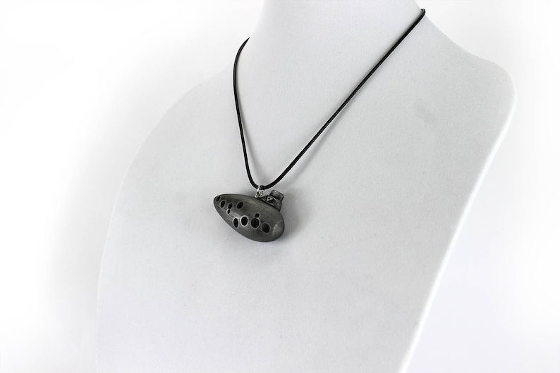 Puede incluir: Un collar con un colgante de ocarina de color plateado y un cord&oacute;n negro. La ocarina tiene un dise&ntilde;o detallado con varios agujeros.