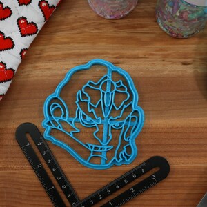 May include: Blue plastic cookie cutter shaped like a cartoon character with a large head and a wide grin. The character has a large nose and eyes.