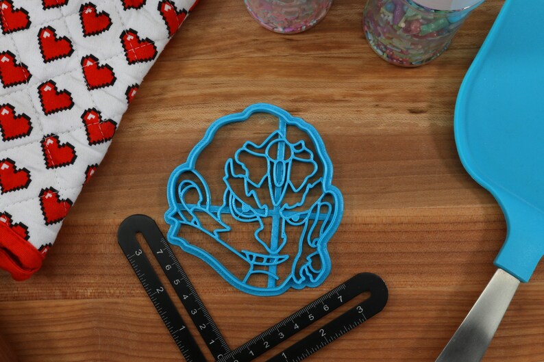 May include: Blue plastic cookie cutter shaped like a cartoon character with a large head and a wide grin. The character has a large nose and eyes.