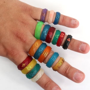 May include: A hand wearing a variety of colorful silicone rings with different symbols and designs. The rings are stacked on each finger.