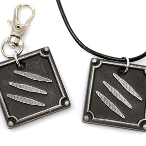 K&ouml;nnte beinhalten: Ein Set aus zwei silberfarbenen Metallanh&auml;ngern mit einem Krallenmuster. Ein Anh&auml;nger ist an einer schwarzen Schnurkette befestigt und der andere an einem silbernen Schl&uuml;sselanh&auml;nger.