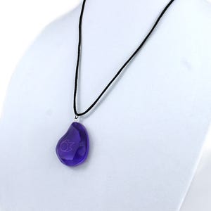 Op de afbeelding: Een ketting met een zwart koord en een druppelvormige hanger van paars glas. De hanger heeft een klein, lichtpaars symbool erin.