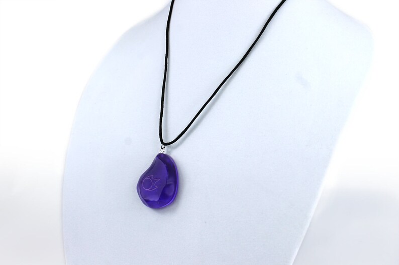 Op de afbeelding: Een ketting met een zwart koord en een druppelvormige hanger van paars glas. De hanger heeft een klein, lichtpaars symbool erin.