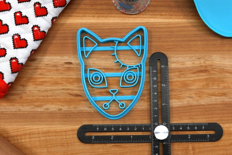 K&ouml;nnte beinhalten: Ein blauer Plastik-Ausstecher in Form eines Katzenkopfes mit gen&auml;hten Augen und einem gestreiften Muster. Der Ausstecher liegt auf einer Holzoberfl&auml;che mit einem Lineal.