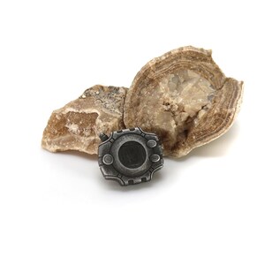 May include: A silver-colored metal pin with a circular design and a black center. The pin is sitting on top of two pieces of brown rock.