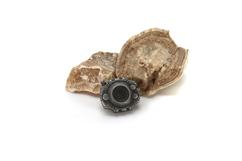 May include: A silver-colored metal pin with a circular design and a black center. The pin is sitting on top of two pieces of brown rock.
