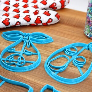 May include: A collection of blue plastic cookie cutters in the shape of a baby carriage and a baby rattle. A red and white oven mitt with heart shapes is in the background, along with a jar of colorful sprinkles.