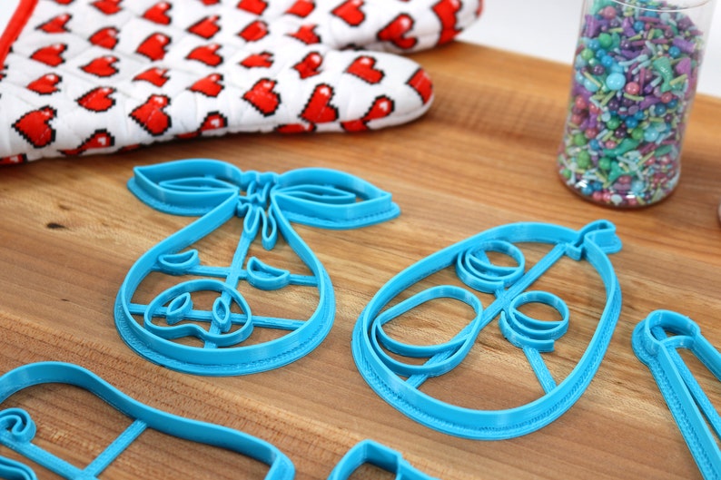 May include: A collection of blue plastic cookie cutters in the shape of a baby carriage and a baby rattle. A red and white oven mitt with heart shapes is in the background, along with a jar of colorful sprinkles.