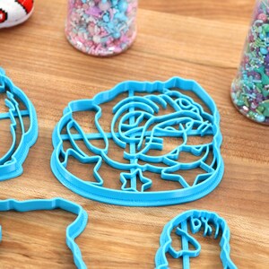 May include: A set of four blue plastic cookie cutters shaped like a pug, a rose, a heart, and a unicorn. The cutters are on a wooden surface.