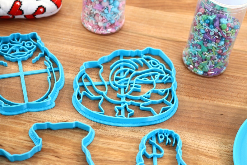 May include: A set of four blue plastic cookie cutters shaped like a pug, a rose, a heart, and a unicorn. The cutters are on a wooden surface.
