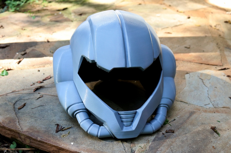 May include: A gray, futuristic-looking helmet with a large visor and intricate details. The helmet is made of a smooth, hard plastic and has a slightly textured surface.