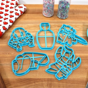 May include: Five blue plastic cookie cutters on a wooden surface. The cutters are shaped like a bottle, a cat, a llama, and two other abstract shapes.