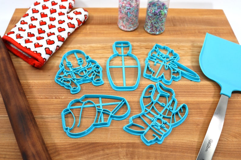 May include: Five blue plastic cookie cutters on a wooden surface. The cutters are shaped like a bottle, a cat, a llama, and two other abstract shapes.