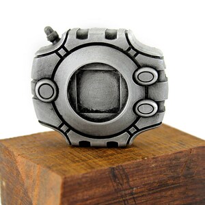 Puede incluir: Una r&eacute;plica de metal plateado de un objeto de videojuego, con un dise&ntilde;o circular y un cuadrado en el centro. La r&eacute;plica se muestra en una base de madera.