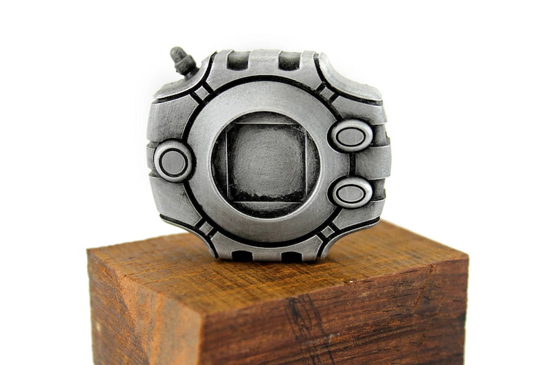 Puede incluir: Una r&eacute;plica de metal plateado de un objeto de videojuego, con un dise&ntilde;o circular y un cuadrado en el centro. La r&eacute;plica se muestra en una base de madera.