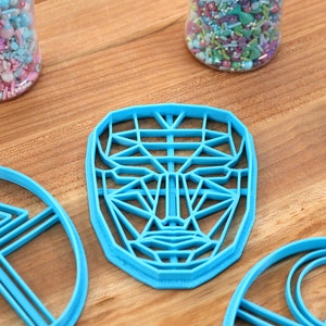 May include: Four blue plastic cookie cutters on a wooden surface. The cutters are shaped like a face, a circle, and two triangles.