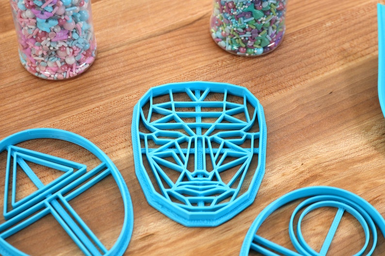 May include: Four blue plastic cookie cutters on a wooden surface. The cutters are shaped like a face, a circle, and two triangles.