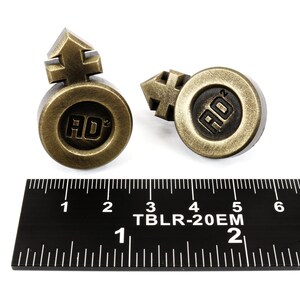May include: Two gold-colored metal buttons with a raised symbol and the letters "AD" in the center. The buttons are about 1 inch in diameter and are sitting on a black ruler.