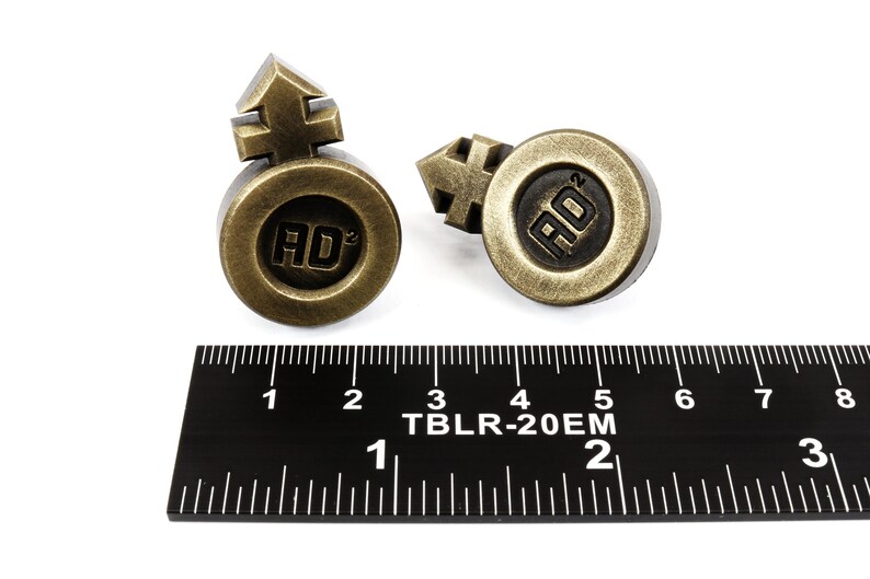 May include: Two gold-colored metal buttons with a raised symbol and the letters "AD" in the center. The buttons are about 1 inch in diameter and are sitting on a black ruler.