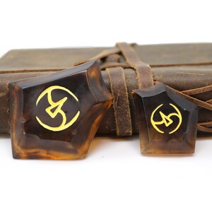 May include: Two brown, geometrically shaped, resin bookmarks with a gold symbol on each. The bookmarks are sitting on top of a brown leather-bound journal.
