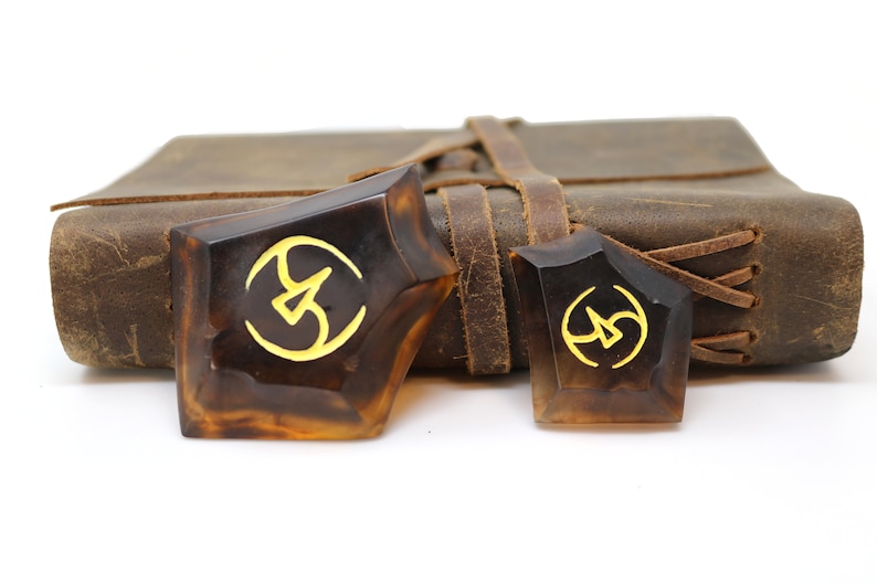 May include: Two brown, geometrically shaped, resin bookmarks with a gold symbol on each. The bookmarks are sitting on top of a brown leather-bound journal.