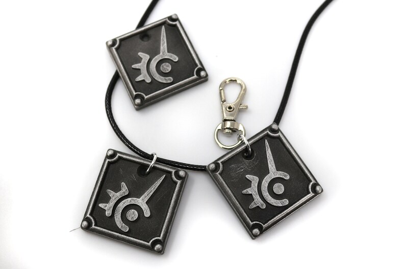 May include: Three silver-colored pendants with a black and white symbol on each. The pendants are attached to a black cord necklace with a silver clasp.