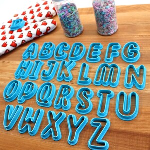 May include: A set of 26 blue plastic alphabet cookie cutters on a wooden surface. The cutters are arranged in alphabetical order.