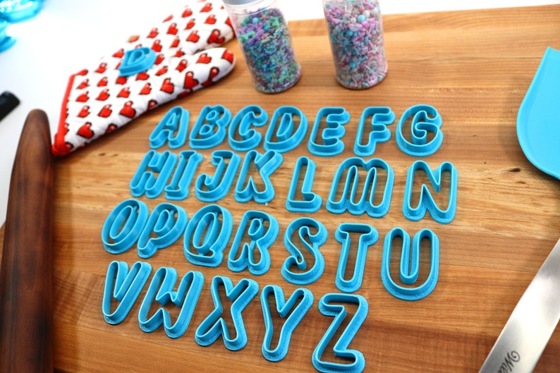 May include: A set of 26 blue plastic alphabet cookie cutters on a wooden surface. The cutters are arranged in alphabetical order.