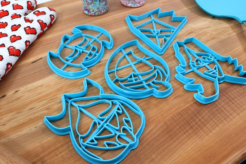 May include: Five blue plastic cookie cutters shaped like cartoon faces. The cutters are on a wooden surface.