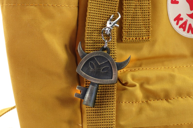 May include: A gold-colored key charm with a shield and horns, attached to a yellow backpack strap.