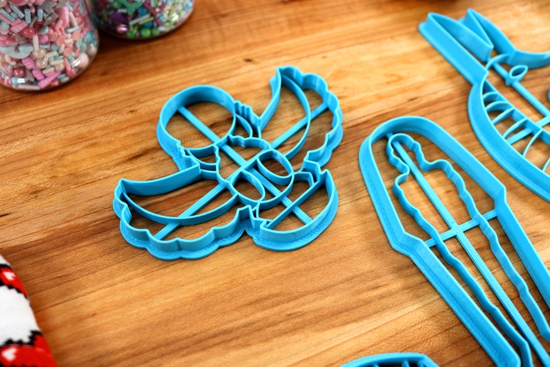 May include: A set of four blue plastic cookie cutters shaped like a person, an angel, a Christmas tree, and a star. The cutters are on a wooden surface.