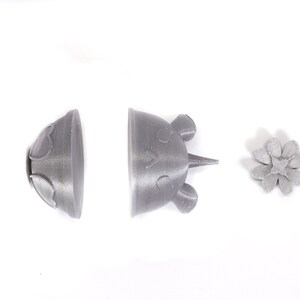 May include: Three 3D printed gray plastic objects: a flower, a dome-shaped object with a heart design, and a crown-shaped object with a face.