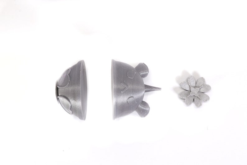 May include: Three 3D printed gray plastic objects: a flower, a dome-shaped object with a heart design, and a crown-shaped object with a face.