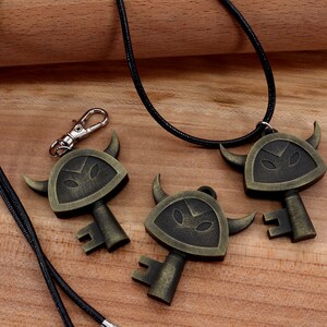 May include: Three gold-toned key-shaped pendants with a black and gold design. The pendants feature a horned, shield-like design with a face in the center. The pendants are on a black cord necklace.