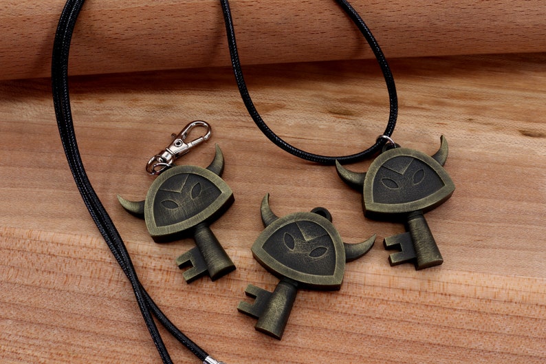 May include: Three gold-toned key-shaped pendants with a black and gold design. The pendants feature a horned, shield-like design with a face in the center. The pendants are on a black cord necklace.