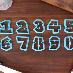 May include: A set of ten blue plastic cookie cutters shaped like the numbers 1 through 10. The cutters are arranged in two rows on a wooden surface.