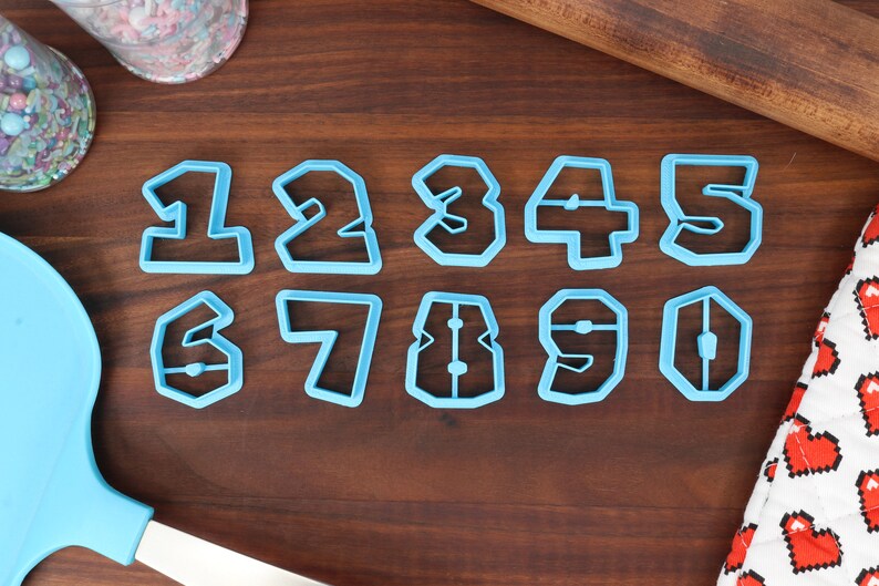 May include: A set of ten blue plastic cookie cutters shaped like the numbers 1 through 10. The cutters are arranged in two rows on a wooden surface.