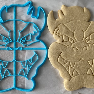 Op de afbeelding: Een blauwe plastic koekjesvormer en een beige koekje, beide in de vorm van een cartoonmonster met een tandenrij en hoorns.