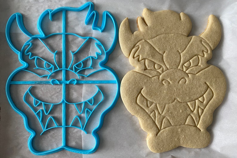 Op de afbeelding: Een blauwe plastic koekjesvormer en een beige koekje, beide in de vorm van een cartoonmonster met een tandenrij en hoorns.