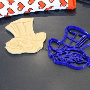 May include: A blue plastic cookie cutter shaped like the Mad Hatter's hat from Alice in Wonderland, next to a baked cookie in the same shape.