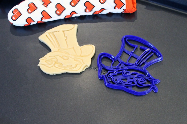 May include: A blue plastic cookie cutter shaped like the Mad Hatter's hat from Alice in Wonderland, next to a baked cookie in the same shape.
